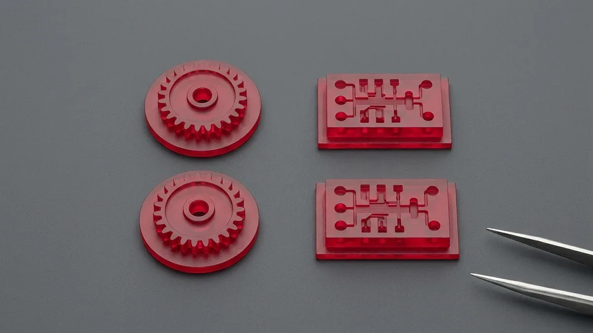 High-detail red castable wax DLP micro-gears alongside optical-clear DLP microfluidic channel chips on black granite
