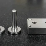 Mirror-polished centerless-ground 440C stainless steel cylindrical dowel pin resting in a hardened 90-degree steel V-block alongside an upright flanged locating pin and a CNC-machined steel housing block with reamed H7 dowel holes on a black granite surface plate.