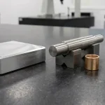 Precision CNC machined aluminum plate, stepped steel shaft on V-blocks, flat-resting bronze bushing, and cross-drilled manifold block on black granite plate