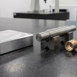 Precision CNC machined aluminum plate, stepped steel shaft on V-blocks, bronze bushing and cross-drilled block resting on black granite metrology plate