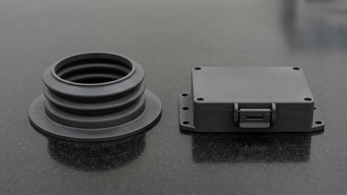Two dark charcoal-grey HP Multi Jet Fusion 3D-printed polymer parts on black granite: flexible TPU 88A convoluted dust boot resting on its wide base flange and rigid PA12 nylon snap-fit electronics housing