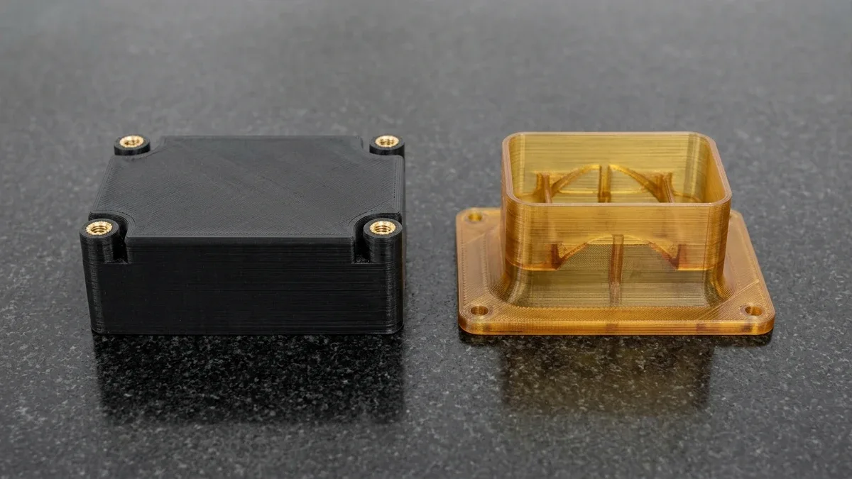 Two industrial FDM thermoplastic parts on black granite: matte-black PC-ABS controller enclosure with heat-set knurled brass inserts and translucent amber ULTEM 9085 flanged air duct