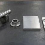 Precision ground 6061-T651 aluminum plate stock blanks and cold-finished stainless steel round bar stock arranged on a workbench beside finished CNC parts
