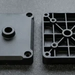 Two matte-black injection-molded engineering thermoplastic structural brackets resting flat on dark granite showing a sink-free cosmetic exterior face and a cored-out underside with uniform wall thickness and stiffening ribs